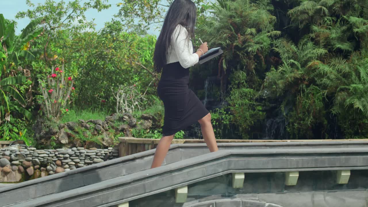 A business-clad girl walking at an outdoor workspace with a pool view.