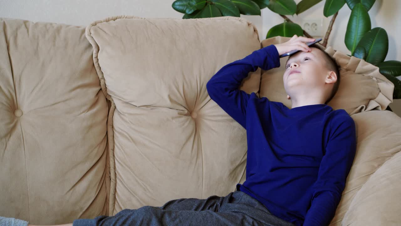 Teenager boy playing on his smartphone becomes angry and throwing gadget at home. Nervous child in casual clothes sitting on the sofa and playing games on a phone.