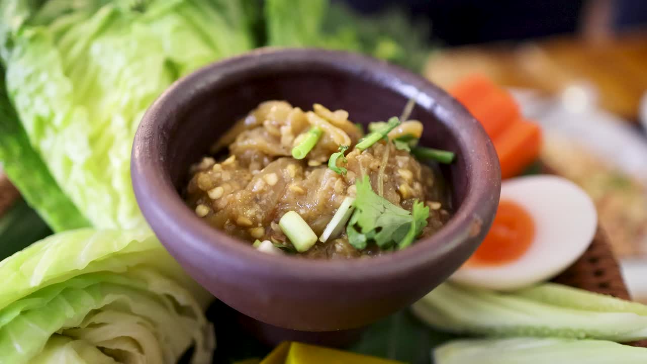 A close-up of a spoon scooping roasted green chili dip from a clay bowl, surrounded by fresh vegetables in natural daylight, shot in Bangkok