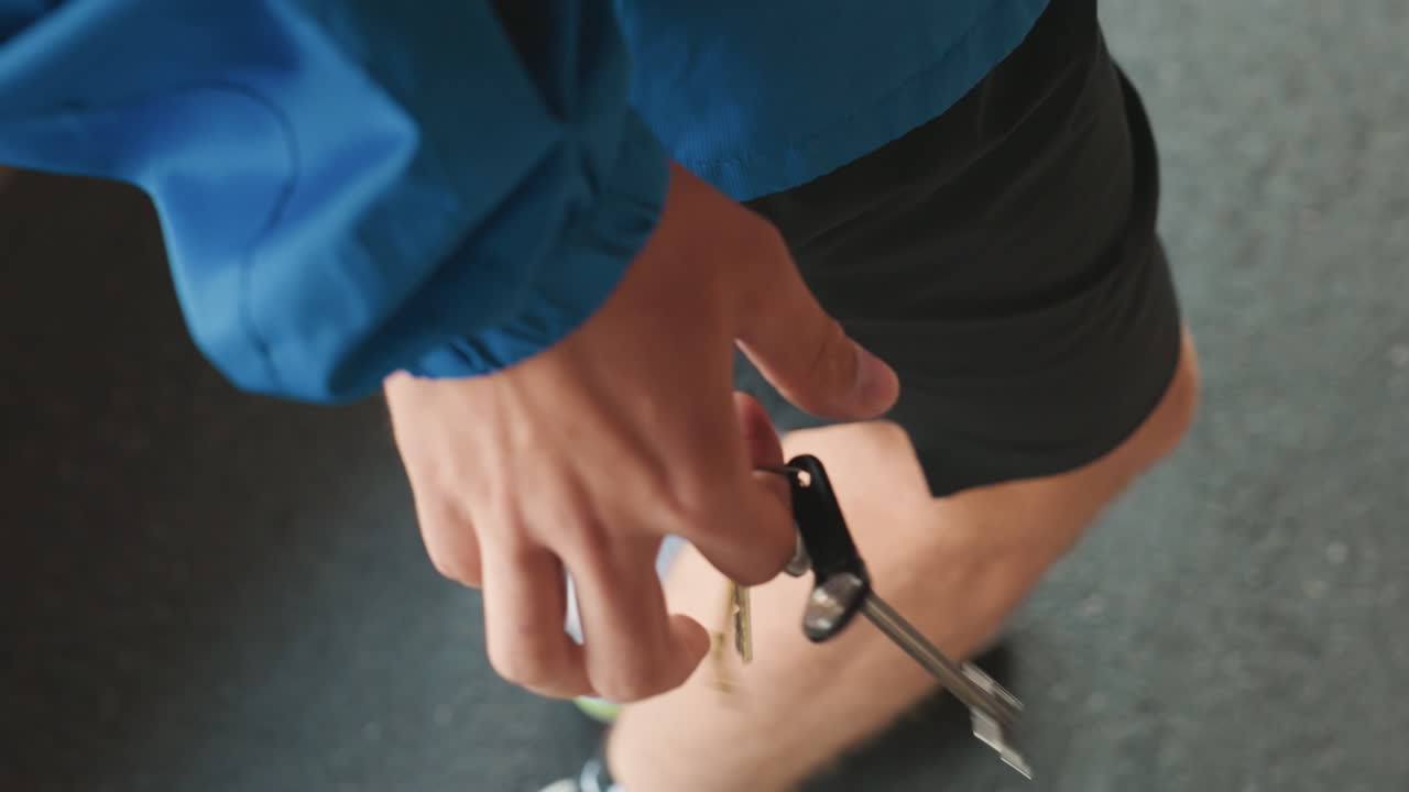 Allenatore che tiene un fischietto e delle chiavi, primo piano della mano a bordo campo bagnato, indossa una giacca blu e pantaloncini neri, scarpe da ginnastica visibili, si prepara a segnalare durante un allenamento di calcio piovoso, pioggerella malinconica, concentrazione intensa