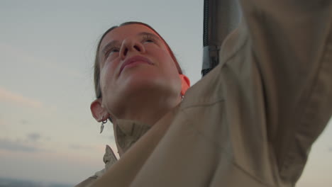 confident woman in outdoor gear raises arm while increasing flame of metal burner during golden hour, warm red light reflecting on face as she focuses upward in calm open-air environment