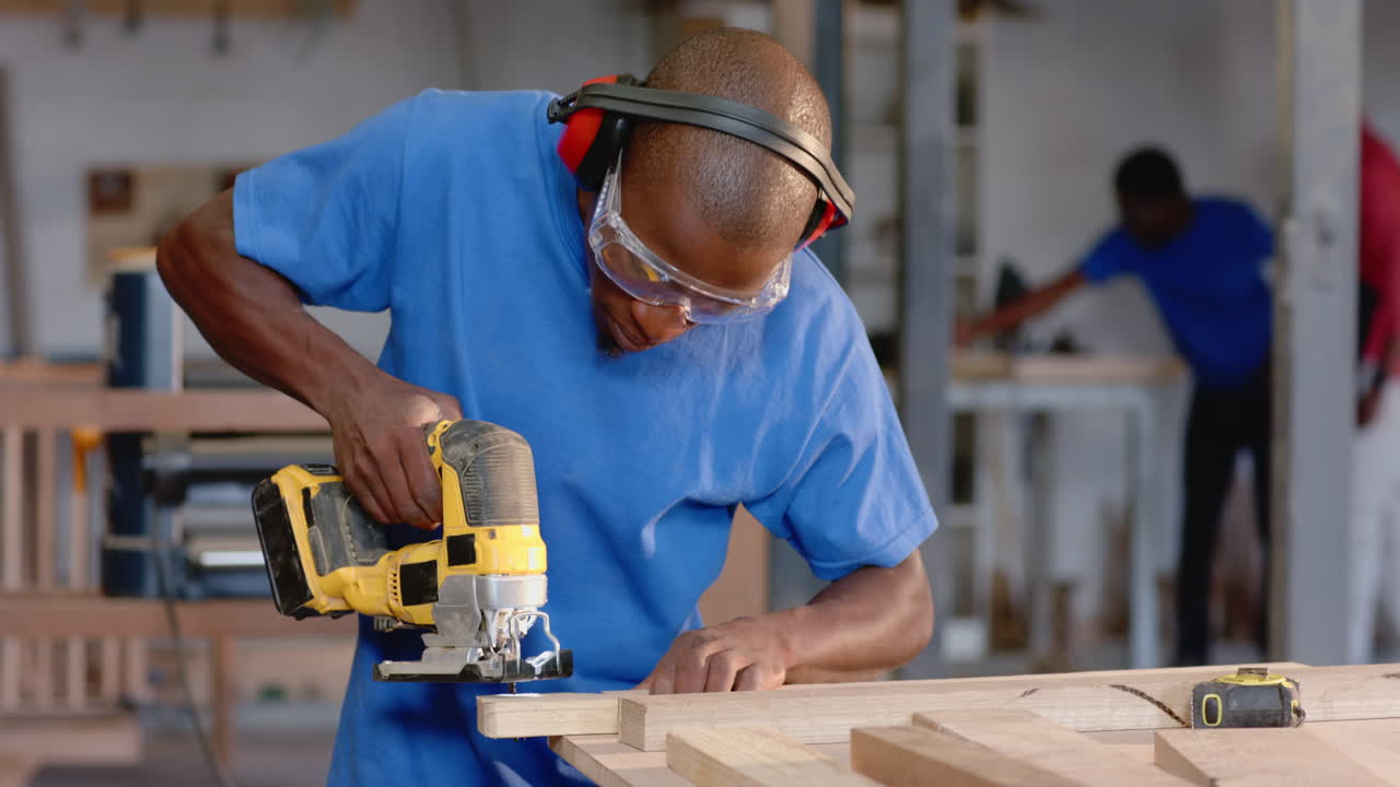 African American coworkers cutting plank in wood shop using electric jigsaw, wearing safety gear