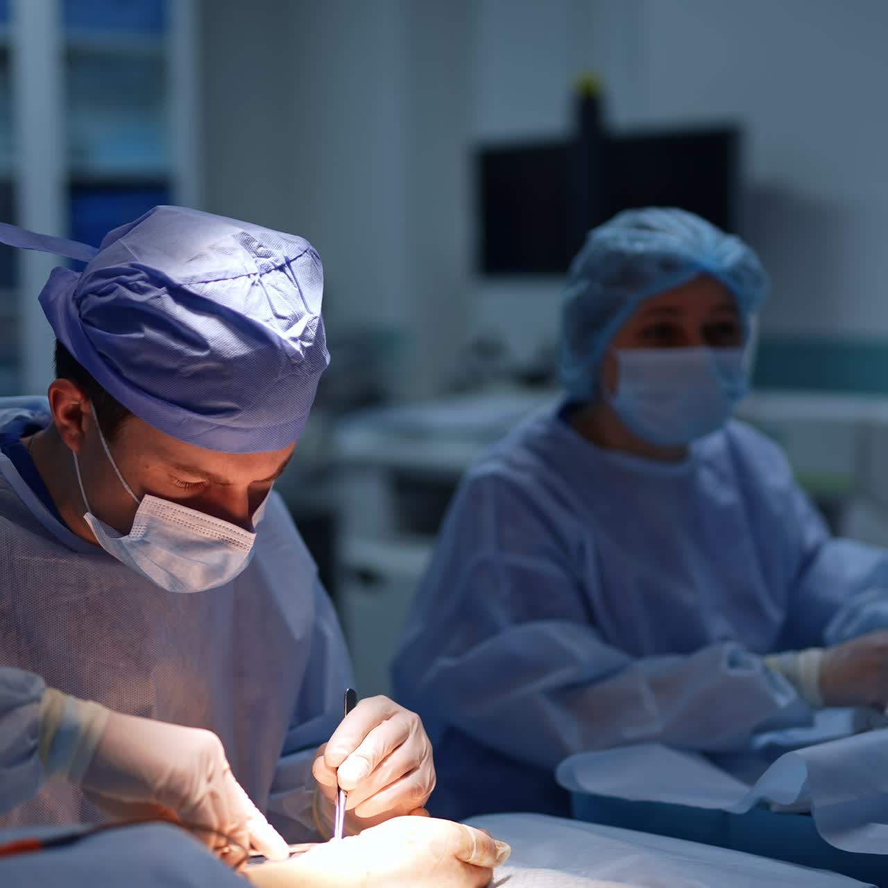 Attentive surgeon carefully using the metal tools performing surgery. Female nurse sitting at backdrop. Blurred background
