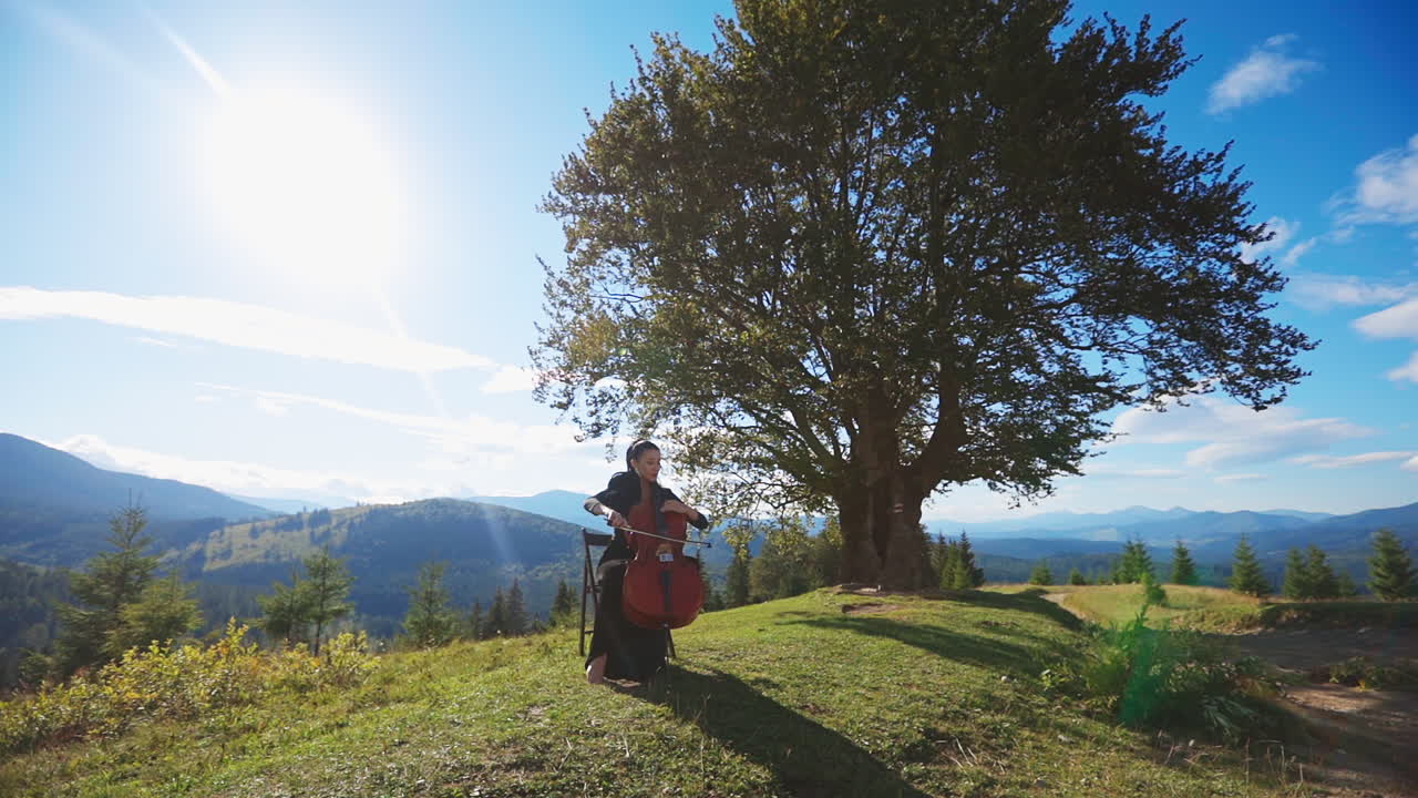 Dark-haired woman in black long dress playing cello. Lady musician making classic music sitting on the chair in the mountainous landscape.