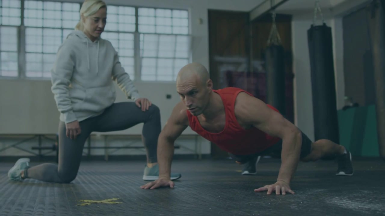 Vertical video: Counting trainer in hoodie cuing man in red top doing push-ups on gym mat for form