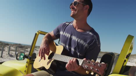 hombre caucásico feliz sentado en un buggy de playa junto al mar tocando la guitarra