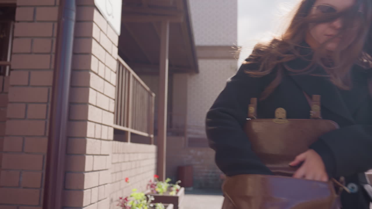 선글라스를 낀 여성이 바람에 머리카락이 날리는 동안 갈색 가방을 정리하고, 햇빛이 현대적인 거리 분위기가 있는 안뜰의 벽돌 난간과 경사로를 스치고, 이른 아침의 차가움, 조용한 외부 보도에 있는 모습이에요.