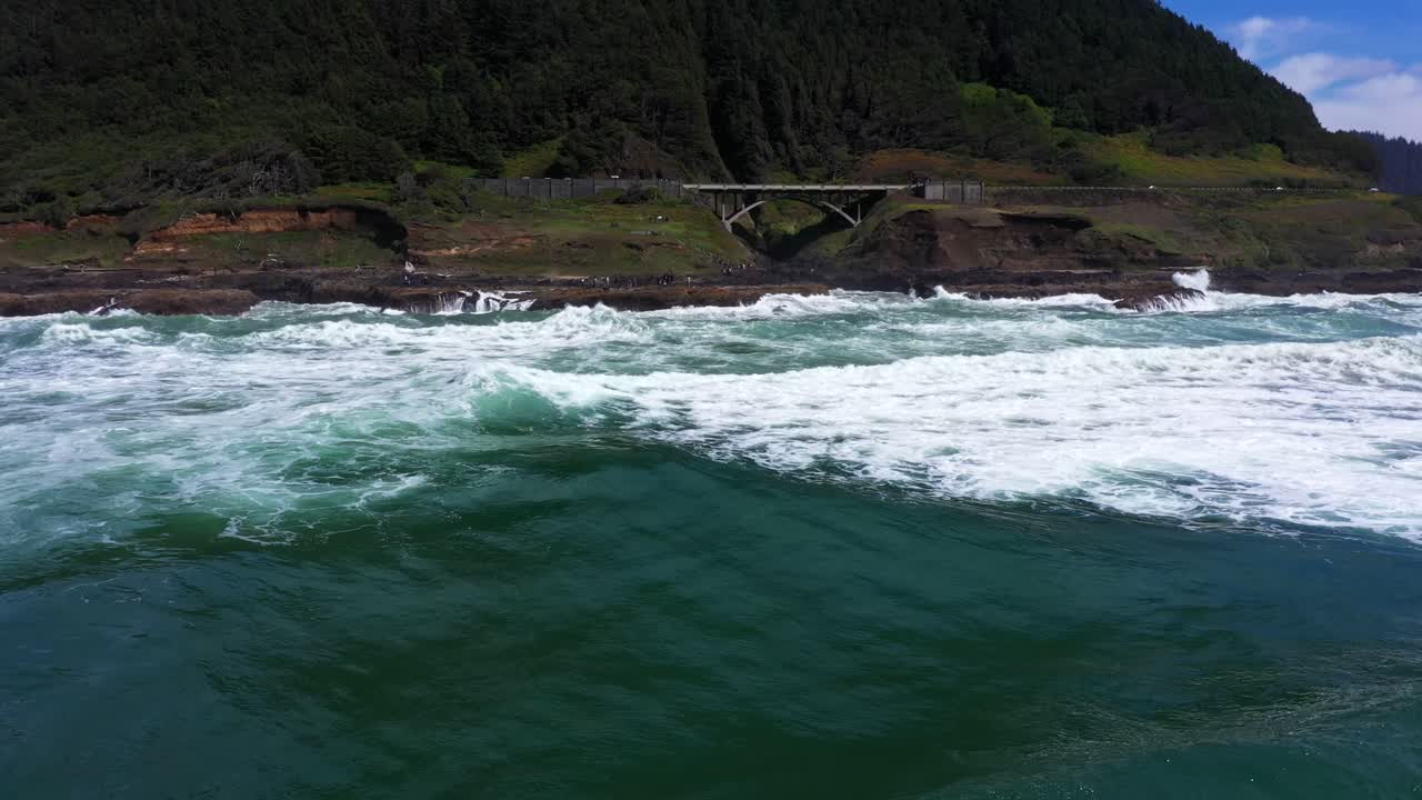 Static aerial shot above the Pacific ocean waves showcasing a hill covered in forest and bridge on the shores of California.