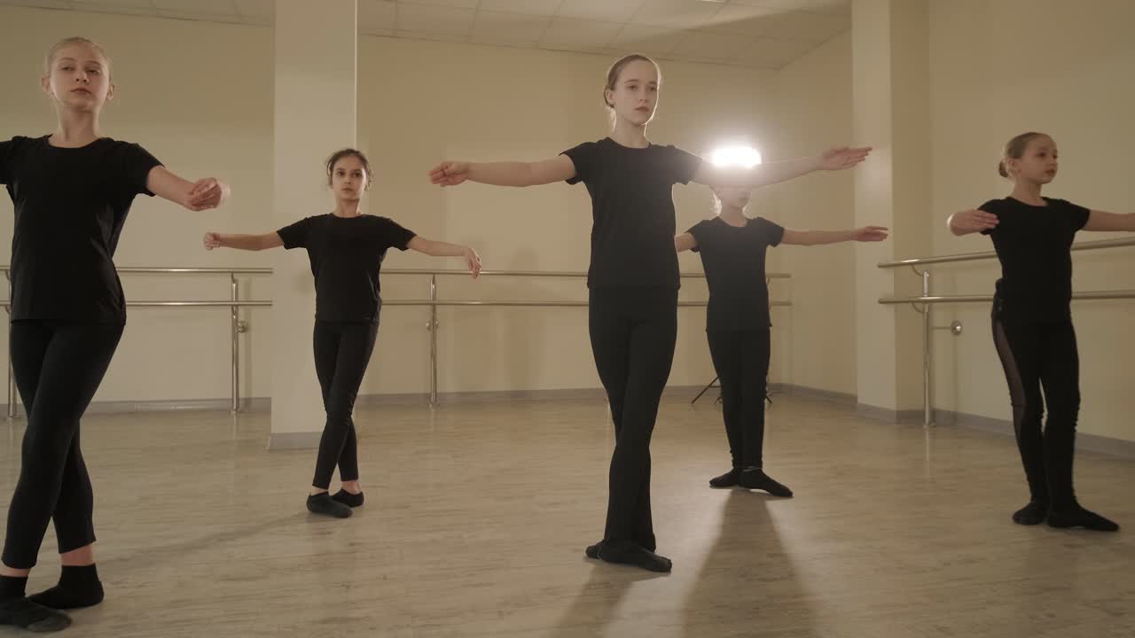 Skupina mladých studentů baletu v černém tanečním oblečení procvičuje pozice v prostorném baletním studiu s dřevěnou podlahou a nástěnnými tyčemi. Soustředěné výrazy a synchronizované pohyby