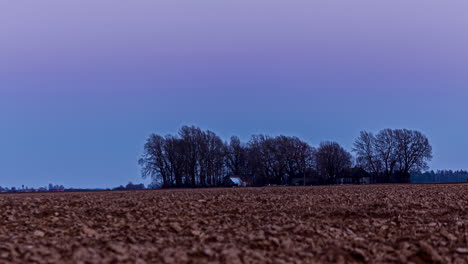 Timelapse-shot-over-agricultural-farmland-soil-during-evening-time-after-sunset-with-the-view-of-trees-in-the-background