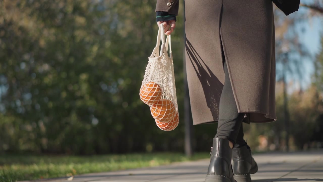Primo piano di una donna che cammina all'aperto, trasportando borse a rete con arance. La donna indossa stivali neri, leggings scuri e un lungo cappotto marrone, mentre tiene una borsa in mano. La luce del sole illumina gli alberi verdi.
