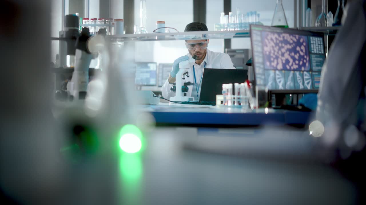 In Bright Medical Science Laboratory: Brilliant Microbiologist Looks Under Microscope Does Analysis of Test Sample. Team of Research Scientists working with High-Tech Equipment and Glassware