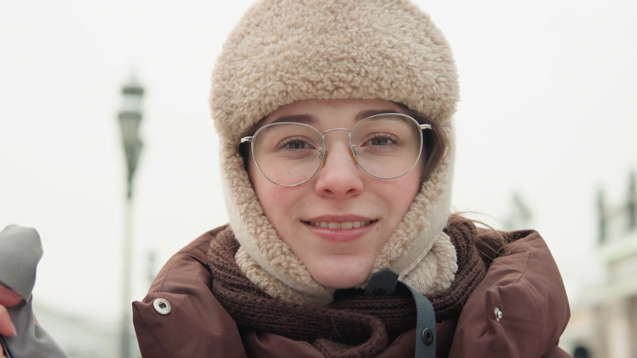 毛皮の帽子、眼鏡、スカーフ、パッド入りコートを着た若い女性が、雪の降る都市の橋の上で電話で話しながら微笑んでいる。彼女の周りにはリラックスした姿勢、冷たい冬の空気、そして柔らかな曇り空の光がある。