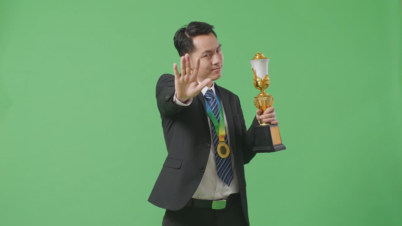 hombre de negocios asiático en traje y corbata con una medalla de oro y trofeo desaprobando sin signos de mano y sonriendo a la cámara en el fondo de la pantalla verde en el estudio