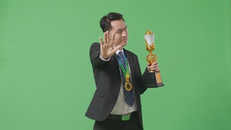 asian business man in a suit and tie with a gold medal and trophy disapproving with no hand sign and smiling to camera on green screen background in the studio