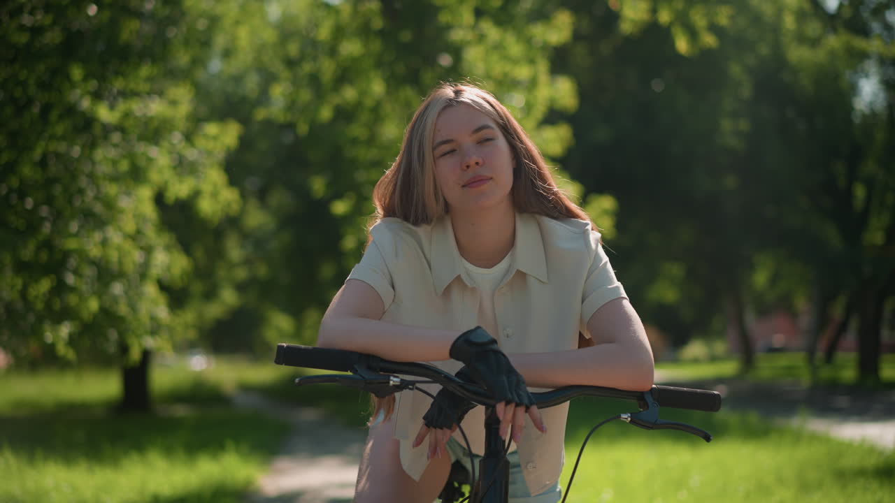 여자는 자전거 잡이에 기대어, 미묘한 미소를 지으며, 빛이 그녀의 얼굴을 비추고, 평온하고 평화로운 표정을 주고, 배경은 활기찬 초록색을 보여줍니다.