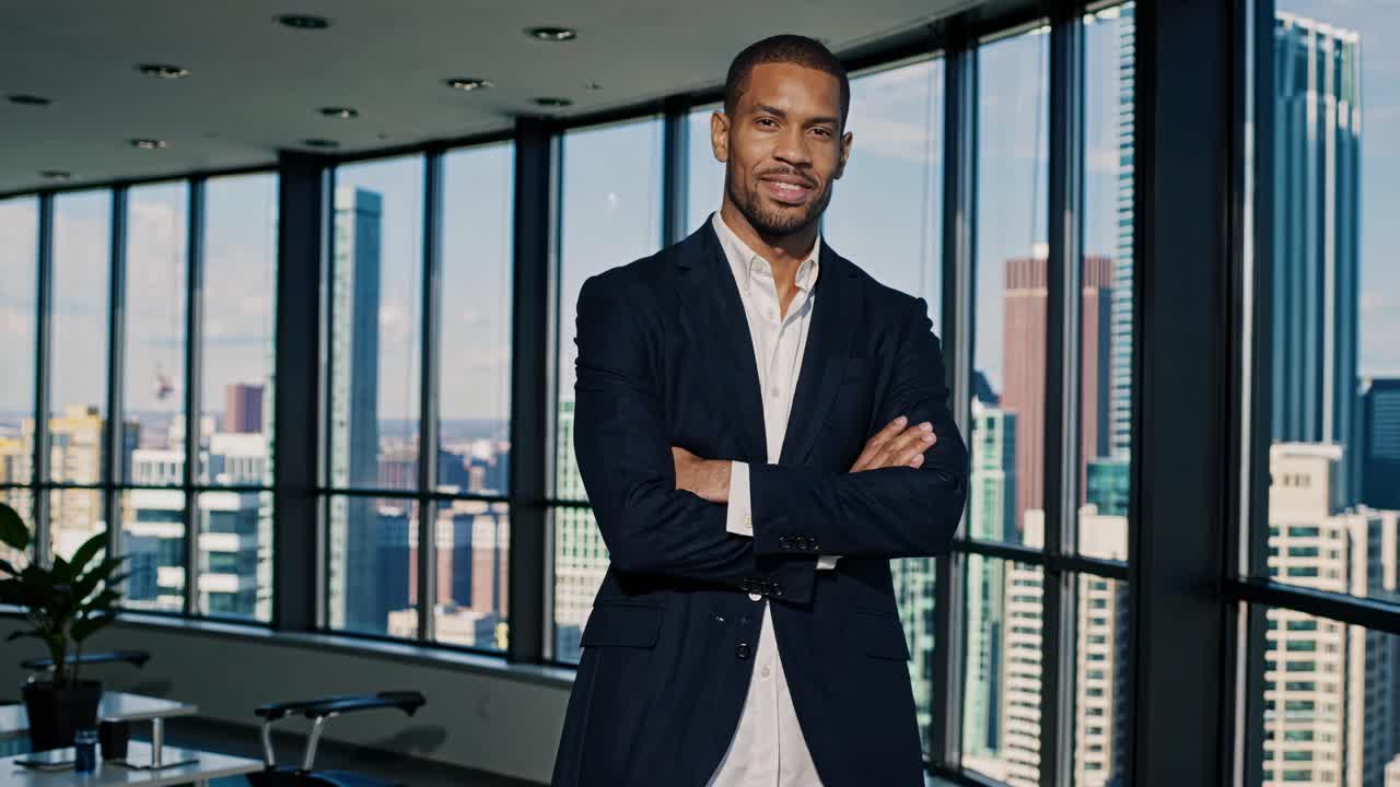 A confident man in a suit stands with arms crossed in a modern office with city views