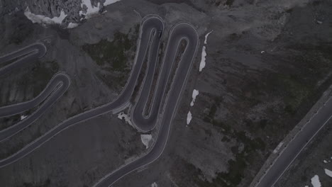 Toma-De-Arriba-Hacia-Abajo-De-Un-Automóvil-Conduciendo-Cuesta-Abajo-En-El-Paso-Stelvio-Italia-En-Un-Día-Gris-Con-Nieve-En-Las-Montañas-En-Una-Carretera-Sinuosa-Y-Faros-Encendidos-Durante-El-Atardecer