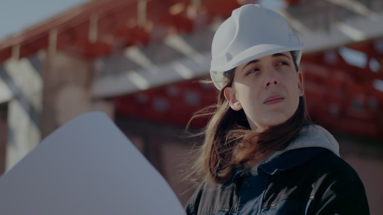 Confident young female architect wearing white hardhat analyzing blueprint at construction site on sunny day