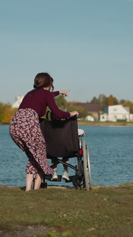 aunt spends time with niece in wheelchair. young woman points finger to house on opposite bank of river. preschooler girl undergoes rehabilitation after injury backside view