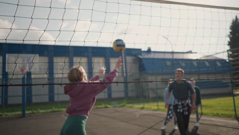 instructor training beginner in volleyball, demonstrating techniques as two female players observe in the background, slightly blurred, outdoor volleyball court with building in the background