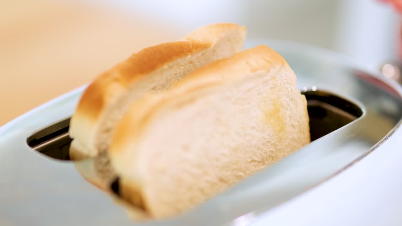 Two slices of white bread rise from a stainless steel toaster in a bright kitchen, captured in close-up with shallow depth of field and natural lighting