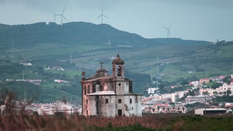 church "ermida de nossa senhora de alcame" on the background of the city of lisbon, green mountains and rotating wind turbines with wheat in the foreground at sunset