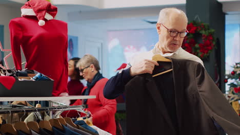 Senior-man-browsing-blazers-in-festive-clothing-store-during-winter-holiday-season,-looking-at-formal-attire-pieces,-doing-Christmas-shopping-spree-in-xmas-decorated-shop