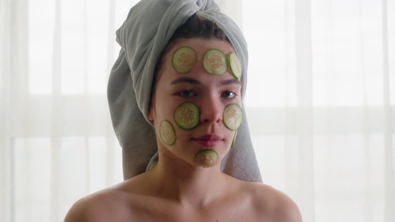 Close-up view of woman with hair wrapped in towel and cucumber slices placed on her face, showing a calm yet determined expression, engaging in relaxing self-care and refreshing skincare moment