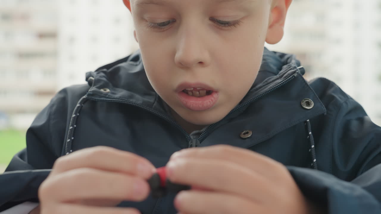 Niño blanco preparándose para la escuela al aire libre, concentrado en un pequeño objeto rojo en sus manos, chaqueta azul marino, edificios de apartamentos al fondo, movimientos cuidadosos de los dedos, expresión pensativa, rutina matutina.