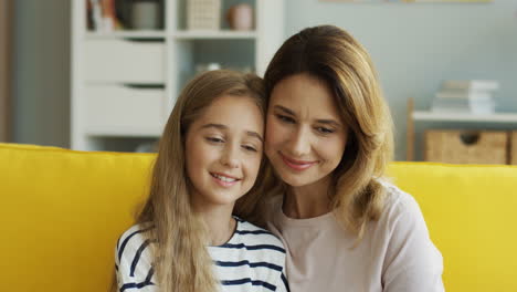 close up view of woman hugging her cute teen daughter while they sitting on couch and watching something in living room