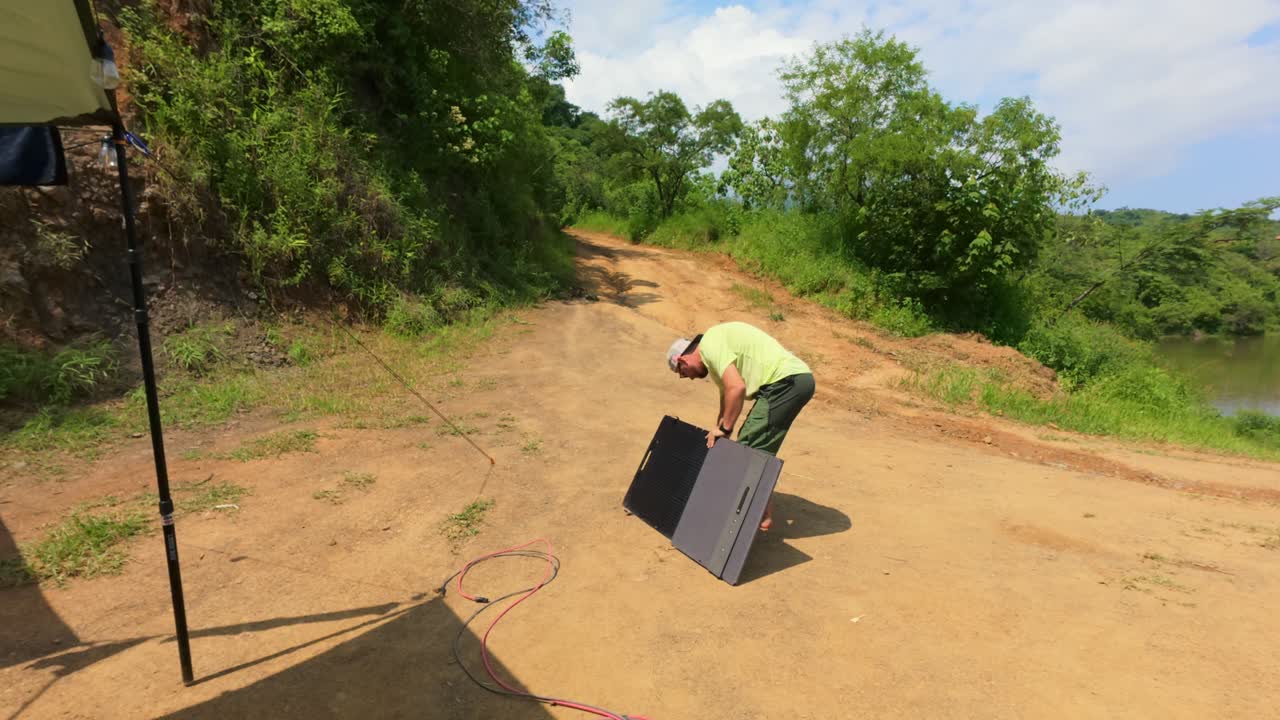 A man sets up a solar panel under bright sunlight in rural Jalisco. Surrounded by lush greenery and a dirt path, this scene captures sustainable energy efforts in natural settings