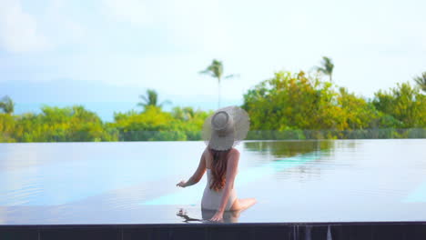 Female-model-in-swimming-suit-and-hat-sitting-at-the-edge-of-pool-touching-water-and-enjoying-the-view-of-tropical-mountains-and-green-trees-on-background,-daytime-slow-motion-back-view