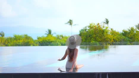 female model in swimming suit and hat sitting at the edge of pool touching water and enjoying the view of tropical mountains and green trees on background, daytime slow-motion back view