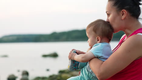 mother is holding a baby in her arms sitting near the river in summer evening. loving mommy kissing her son in head. blurred backdrop.