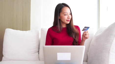 A-pretty-young-Asian-woman-sitting-on-an-overstuffed-couch-holds-a-credit-card-in-one-hand-while-she-inputs-the-number-into-a-laptop-balanced-on-her-lap