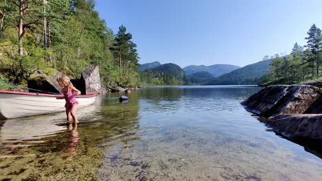 Niño-Rubio-Con-Traje-De-Baño-Rosa-Vadeando-Y-Jugando-Con-Un-Bote-En-La-Playa---Bahía-Acogedora-En-Un-Lago-De-Agua-Dulce-Durante-El-Verano-En-Noruega---Noruega-Occidental-Estática