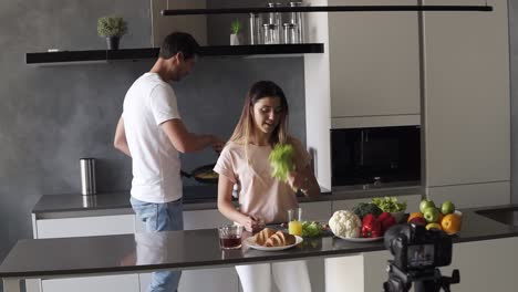 Young-attractive-couple-bloggers-in-casual-clothes-shooting-video-food-blog-about-their-daily-lifestyle,-cooking-simle-breakfast-on-camera-in-the-modern-kitchen