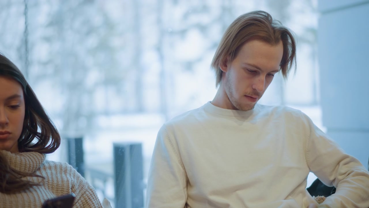 Close up of two young adults sitting side by side indoors each focused on their smartphones. woman wears knitted sweater and man in plain white clothing both maintain steady positions