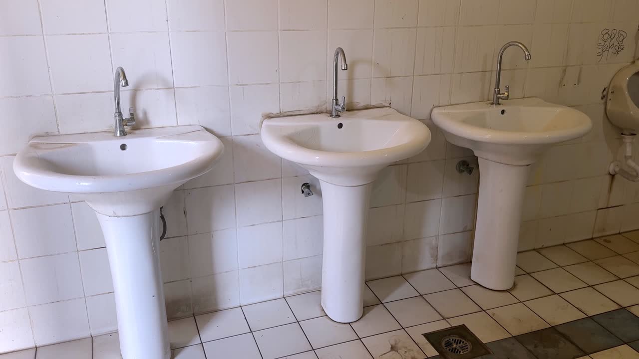 Vista statica di tre lavabi a colonna bianchi allineati contro una parete piastrellata e macchiata in un bagno pubblico interno, che mostra rubinetti, scarico a pavimento e dettagli usurati di un bagno di servizio