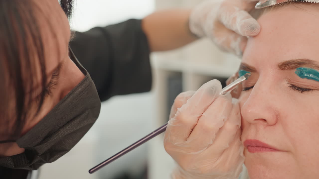 Closeup woman receiving teal eyeshadow application in studio with makeup artist wearing gloves, brush applying shimmer to eyelid, calm expression and precise strokes, cosmetic palette in background