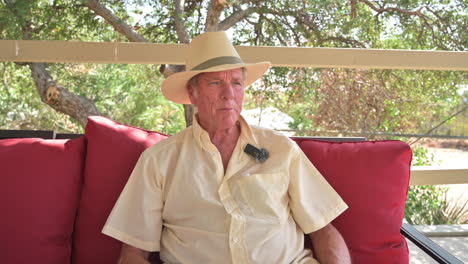 Portrait-of-good-looking-man-70s-being-interviewed,-wearing-wireless-mic,-sitting-on-patio-chair-outdoors