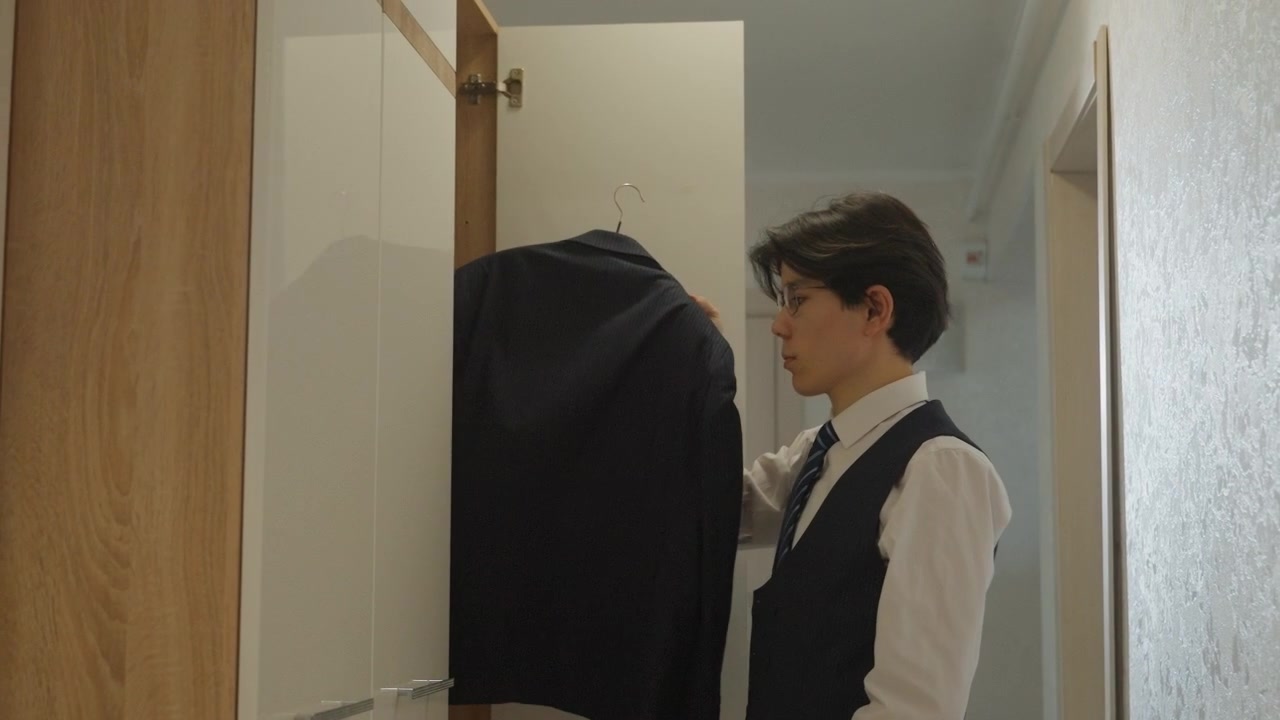 Giovane uomo asiatico nel corridoio prende giacca, camicia bianca e gilet blu scuro, apre l'armadio, solleva la giacca dell'abito, controlla la vestibilità, aggiusta la postura, una calma luce mattutina filtra nel corridoio di un appartamento minimalista.