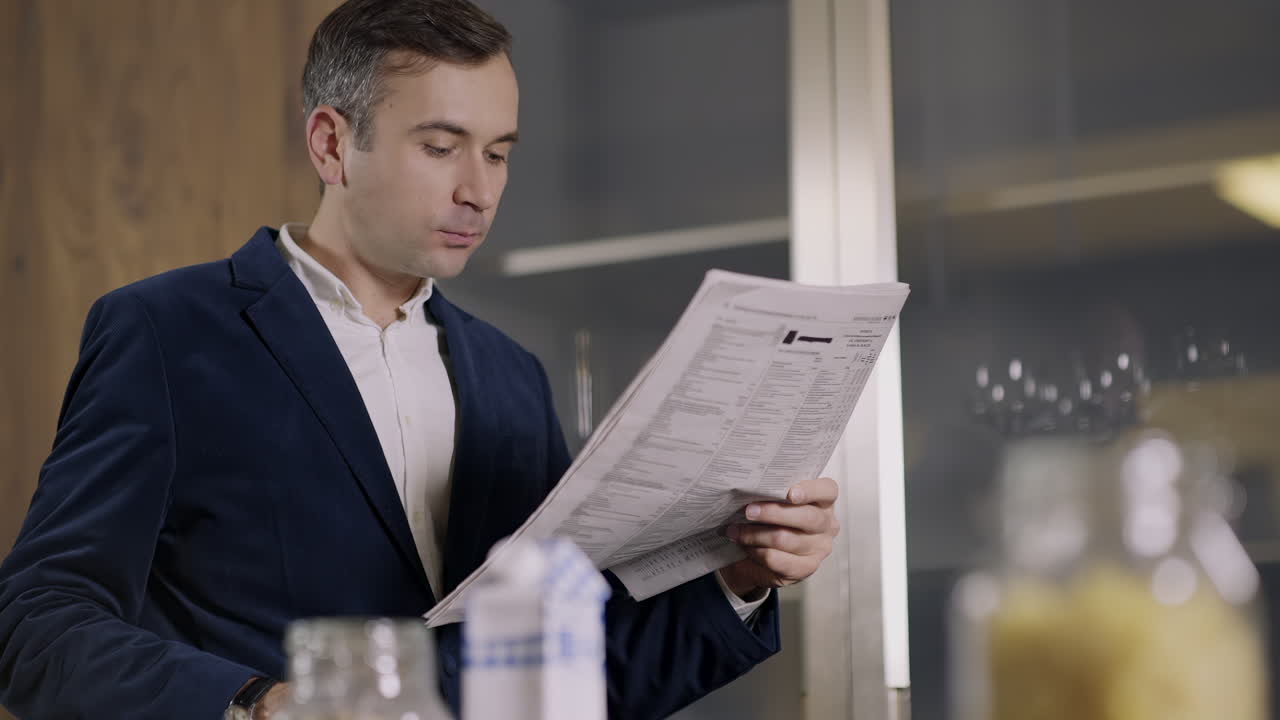 A man sits in a stylish kitchen, savoring his meal with one hand while holding a newspaper with the other. Morning light fills the space, creating a cozy atmosphere