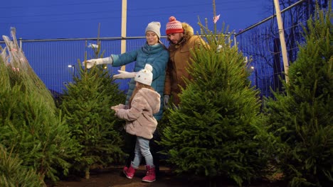 Glückliche-Familie-Kauft-Weihnachtsbaum-Auf-Dem-Markt.Familie,-Winterferien-Und-Menschenkonzept-–-Glückliche-Mutter,-Vater-Und-Kleine-Tochter-Kaufen-Weihnachtsbaum-Auf-Dem-Straßenmarkt-Am-Abend-Bei-Fallendem-Schnee