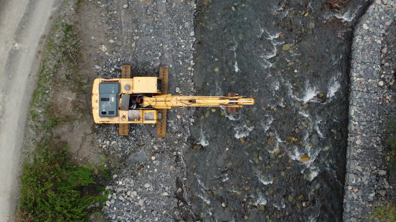 Top-down drone view of an industrial yellow excavator standing on rocky riverbed during construction