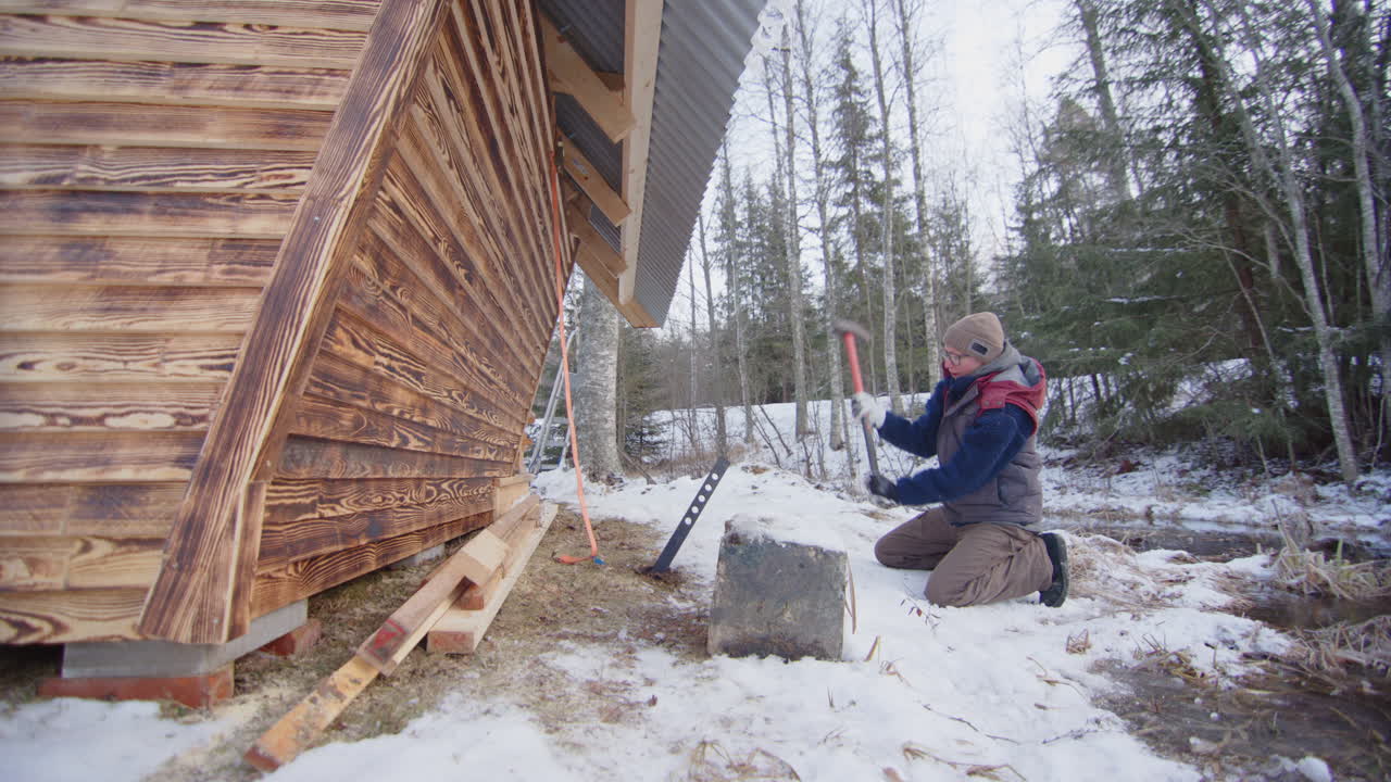 una mujer golpea un poste de acero en el suelo para anclar una pequeña cabaña de madera en una tormenta de invierno