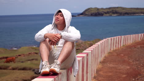 Un-Joven-Asiático-Sentado-En-Un-Banco-Con-Un-Impresionante-Paisaje-Marino-En-El-Fondo-Durante-Las-Vacaciones-De-Verano.