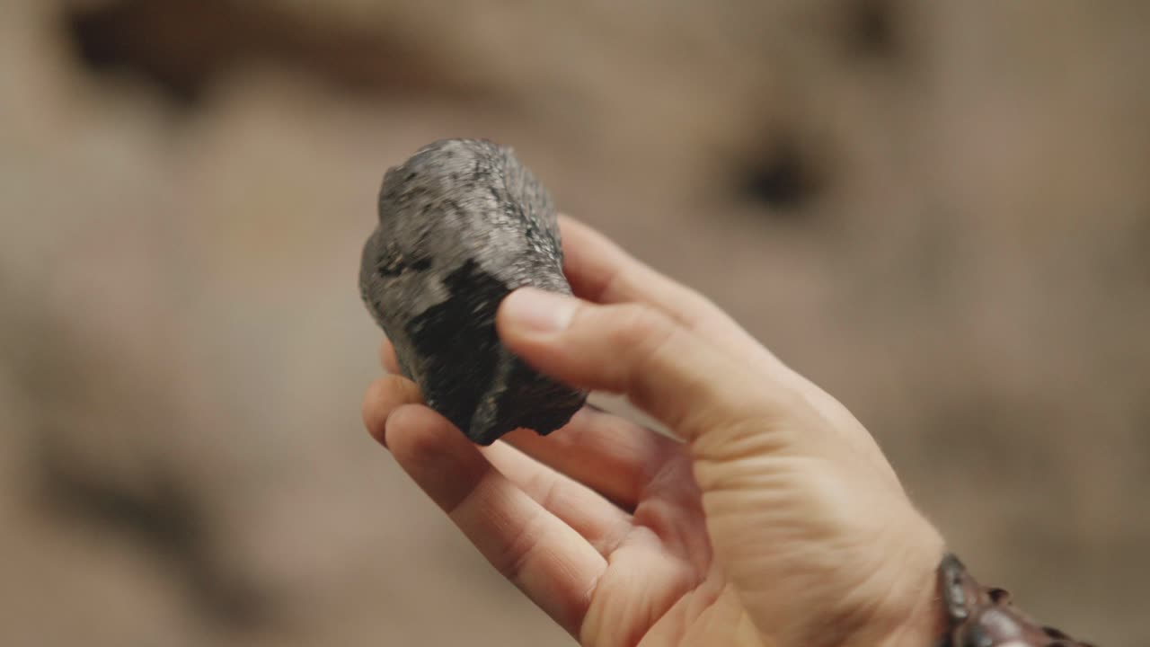 A close-up shot of a hand inspecting a raw, black mineral. Historical, fantasy, or geology concept, showing the discovery of a valuable resource like coal or obsidian