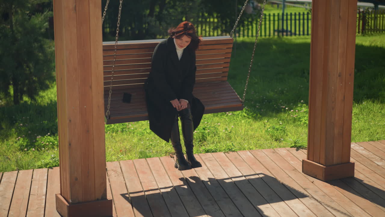 una dama con un elegante abrigo negro y botas se sienta en un columpio de madera en un parque iluminado por el sol, mirando serenamente a la distancia, el teléfono en la silla, la valla de hierro en el fondo